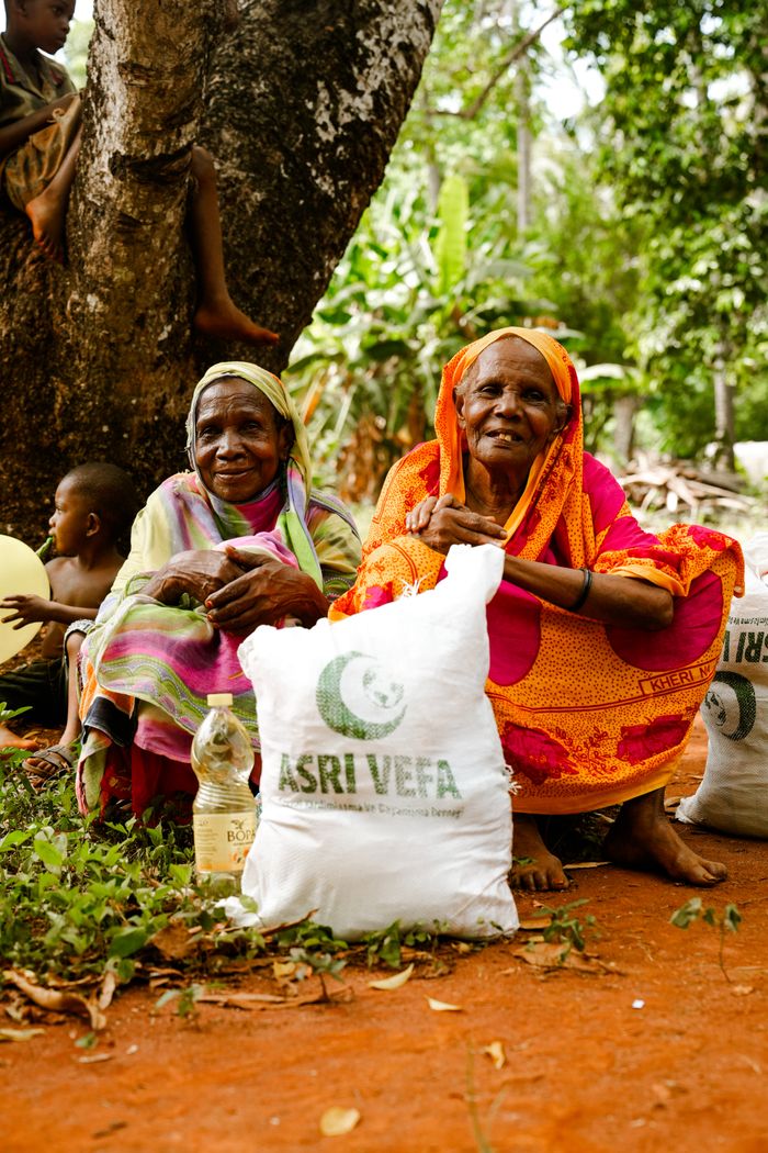 services-04 Two elderly women hold aid packages in a rural outdoor setting, sitting on red soil.